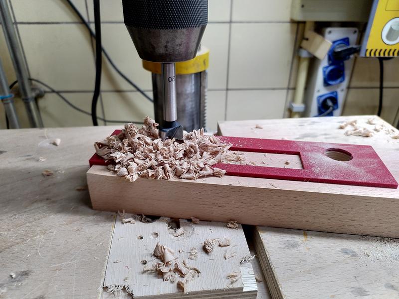 A piece of beech (fagus sylvatica) on the table of a pillar drill.
There's a red PLA 3d printed template on the top of the wood, a pile of
wood chips on the left, and a 20mm forstner bit in the chuck of the drill.