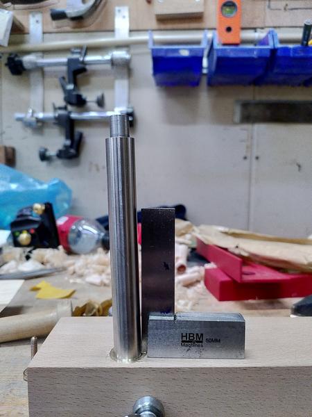A polished steel bar sticks up out of a piece of beech (fagus
sylvatica). A machinests square is resting next to the bar, showing a
pronounced lean to the bar. In the background is an exceptionally messy
workbench.